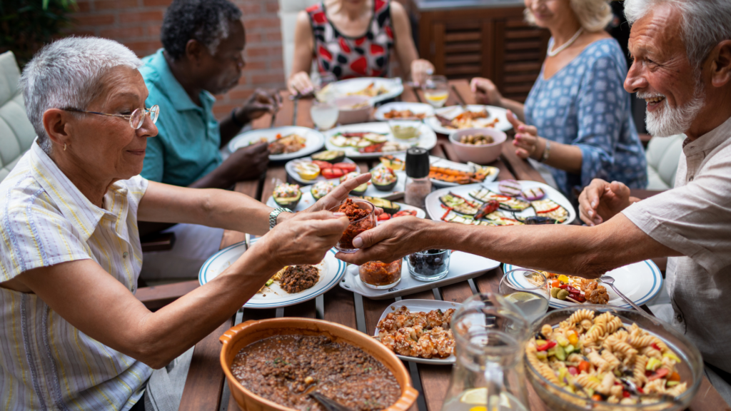 Un gruppo di persone anziane di diverse origini si riunisce attorno a un tavolo all'aperto, condividendo un pasto colorato e variegato. Piatti con verdure grigliate, insalate di pasta, legumi e altre pietanze salutari riempiono la tavola. Una donna con i capelli corti grigi e occhiali passa un piccolo contenitore di crema o salsa rossa a un uomo con la barba bianca, entrambi sorridenti. L'atmosfera è conviviale e rilassata, con persone che si scambiano cibo e conversano piacevolmente.